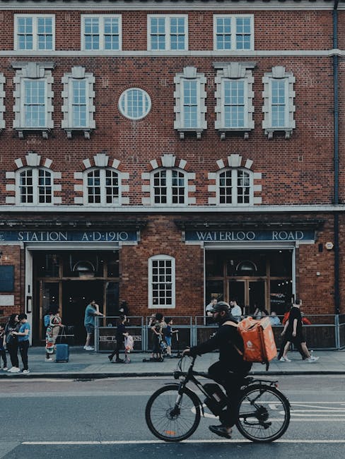 A person riding a bicycle with a black frame, wearing a black jacket and a grey cap, passes by a bustling street scene in front of a red brick building with white framed windows and a sign indicating