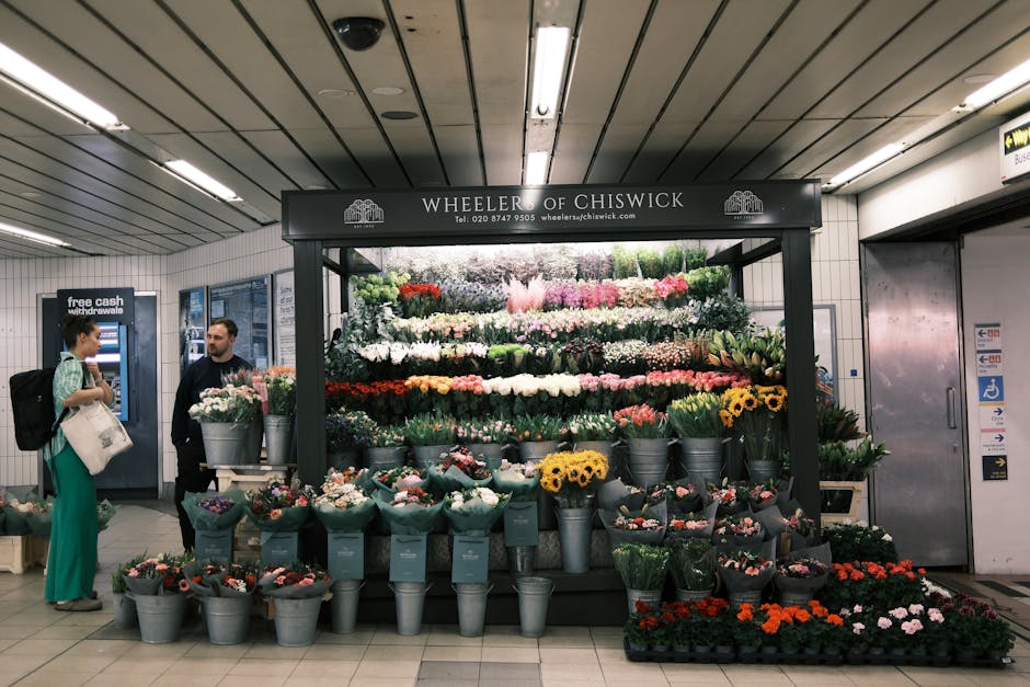 A floral display at Wheeler of Chiswick in an underground transit station features an assortment of fresh flowers arranged in metal buckets. The bouquet includes bright yellow sunflowers, pink roses,