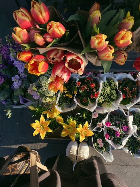 A top-down view of a flower display featuring bouquets of tulips in shades of yellow and red with orange accents, arranged on a dark surface. Surrounding the tulips are smaller potted plants and flowe