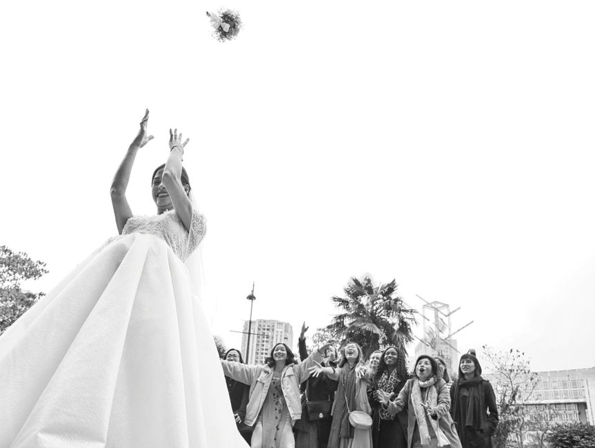 A woman wearing a white wedding gown with lace details on the bodice and a full tulle skirt is joyfully tossing a bouquet of fresh flowers into the air during an outdoor celebration. Her arms are rais
