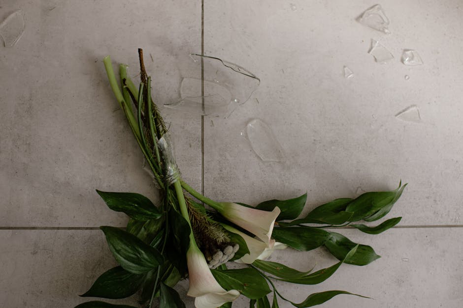 A fallen bouquet of fresh white calla lilies with long green leaves and stems, wrapped loosely in clear plastic, lies on a light-colored tiled floor. The bouquet appears to have been accidentally drop