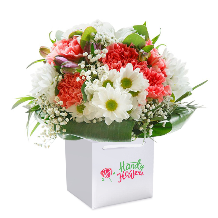 A beautifully abundant floral bouquet sits elegantly within a pristine white, square-shaped gift bag, its freshness illuminated by soft, natural light. Bursting with locally sourced coral, peach, and scarlet carnations, each flower reveals an intricate maze of ruffled petals, conveying a sense of both energy and delicate craftsmanship. Crisp white daisies, defined by their unmistakable citrus-green centers, provide a refreshing juxtaposition-like a breath of fresh air found along the paths of Southwark Park. Threads of feathery white gypsophila (baby's breath) swirl gently among the larger blooms, introducing a gossamer softness throughout the composition. At one edge, a few slender, purple-blushed buds-perhaps an early sign of seasonal alstroemeria-add depth and a hint of mystery, much like the quiet charm of the historic Rotherhithe docks. The bouquet is encircled by a robust array of lustrous, deep-green leaves, each shining with vitality and forming a supportive framework akin to a natural collar. The squared white bag is thoughtfully designed for secure delivery, with sturdy ribbon handles folded neatly at the top-ideal for carrying to a loved one's door. Clear yet understated branding signifies a trusted local service, resonating with Rotherhithe's close-knit feel. Crafted for moments of joy-whether birthdays at Canada Water or spontaneous gestures along Salter Road-the bouquet's perfectly balanced shape and fresh palette reflect the artistry of local florists and the authentic spirit of Rotherhithe's vibrant riverfront community.
